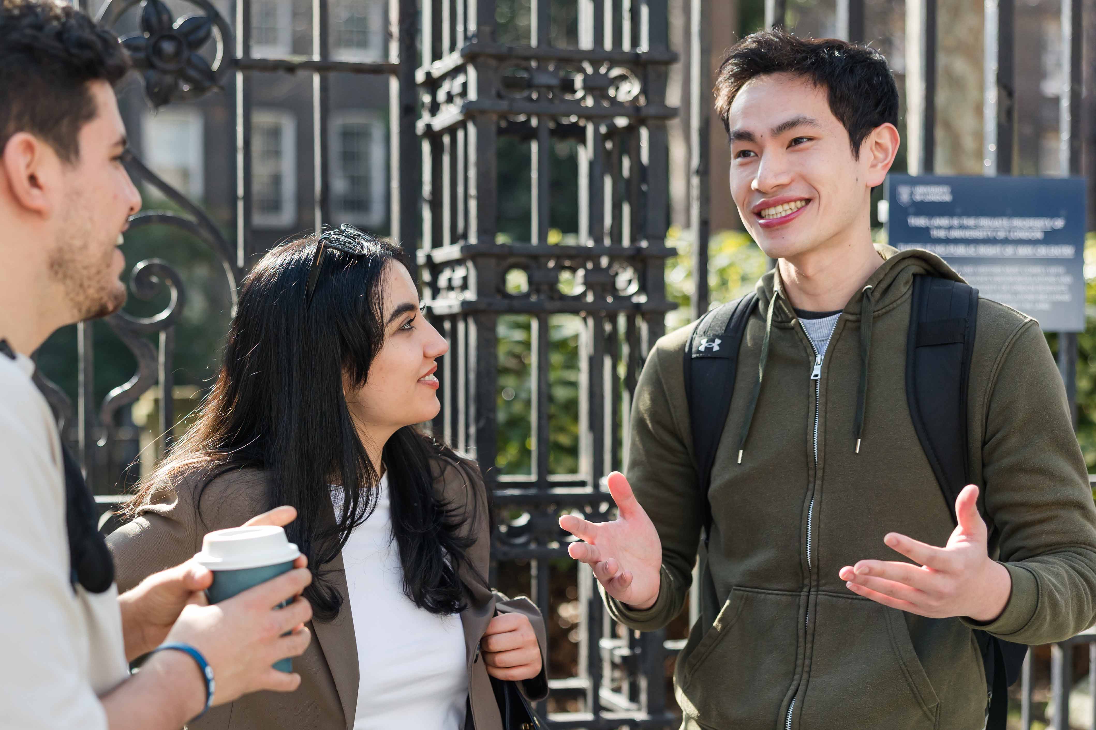 Three young adult students standing outside near university gates in London chatting while holding coffee.