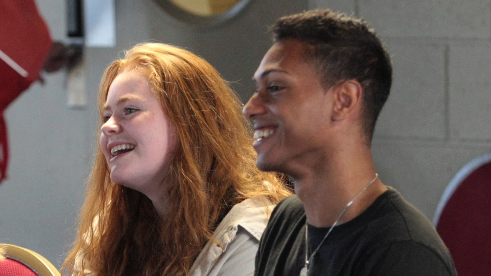 A young man with short dark hair and a young woman with long red hair smiling and laughing together while sitting in an audience.