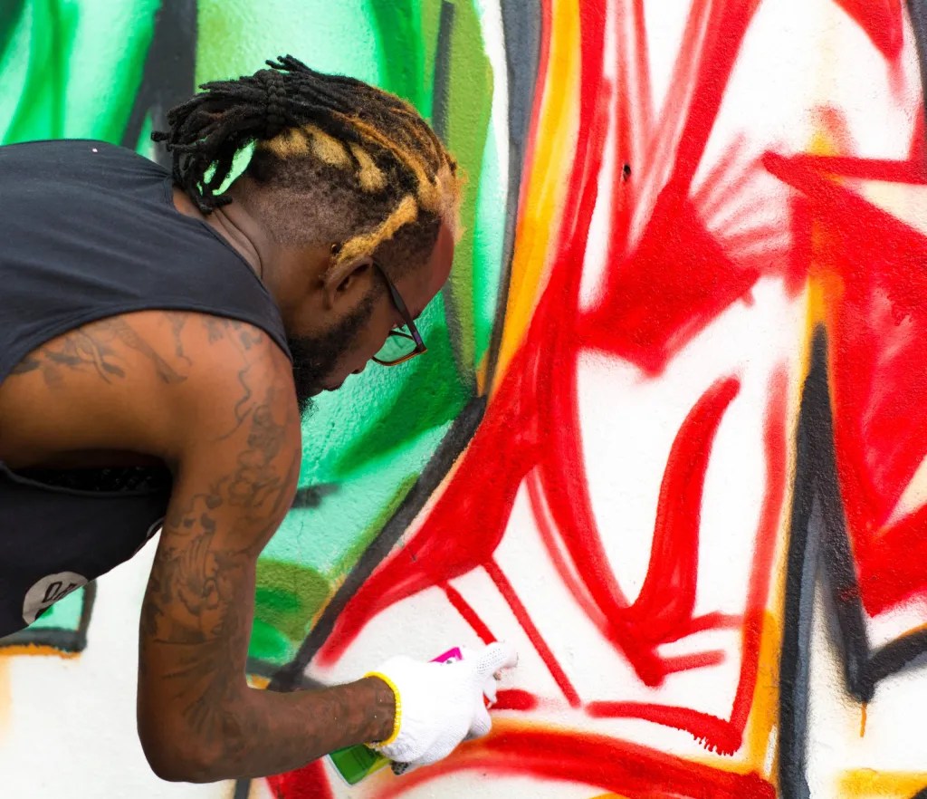 A man with blonde-tipped dreadlocks wearing white gloves while spray-painting a colorful graffiti mural with red and green paint.