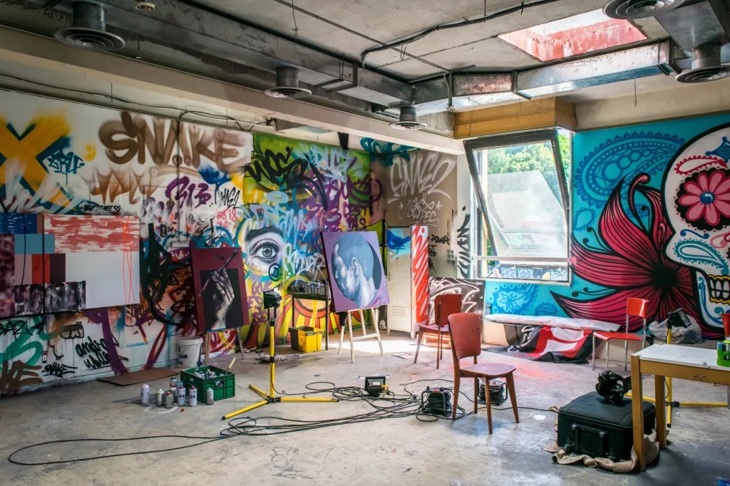 An indoor creative art studio space featuring extensive graffiti wall murals, paintings on easels, and wooden chairs.