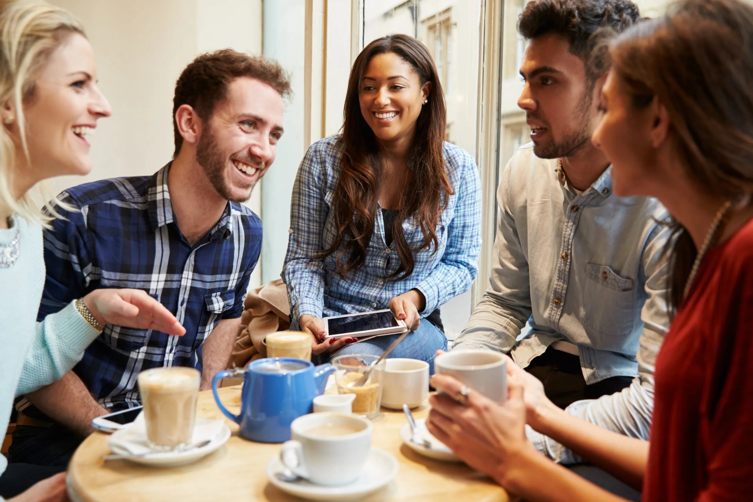 A diverse group of five young adult friends smiling, talking, and drinking coffee together around a small table at a café.
