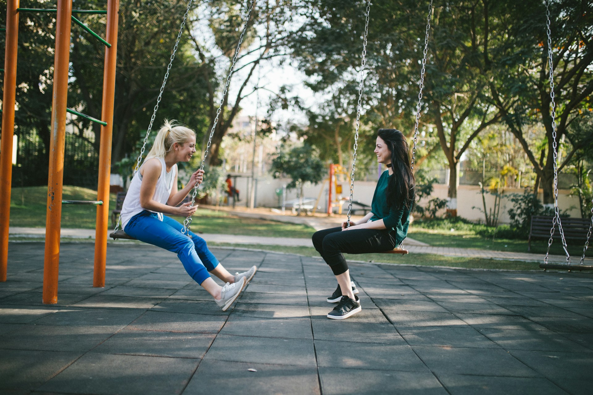 Two friends sitting on swings at an outdoor park, facing each other and laughing.