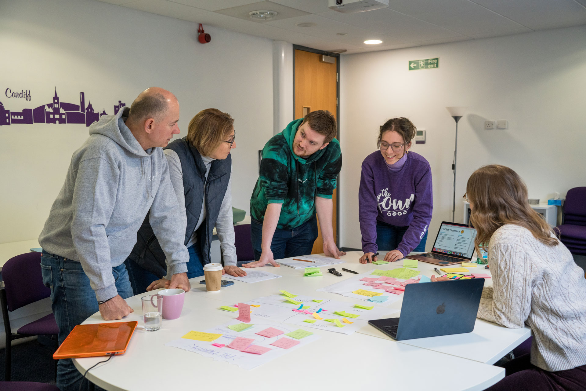 A group of five people collaborating around a table filled with laptops, printed papers, and colorful sticky notes during a NextSteps Greenhouse event. They are actively discussing and brainstorming digital outreach journeys in a bright meeting room.