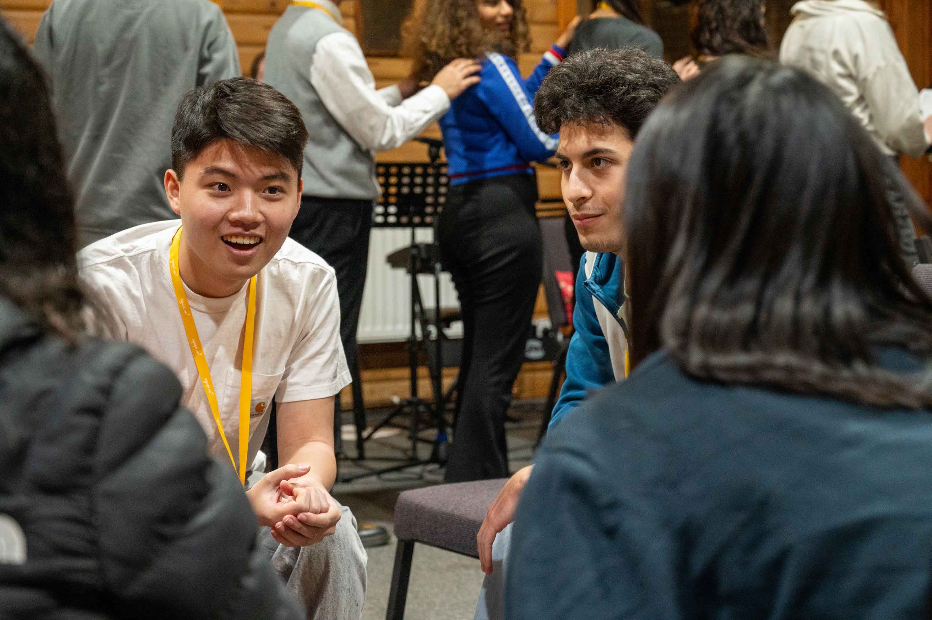 A diverse group of young adults sitting in a circle engaged in a friendly conversation. A smiling young man in a white t-shirt and yellow lanyard is speaking to the group, while others listen attentively.