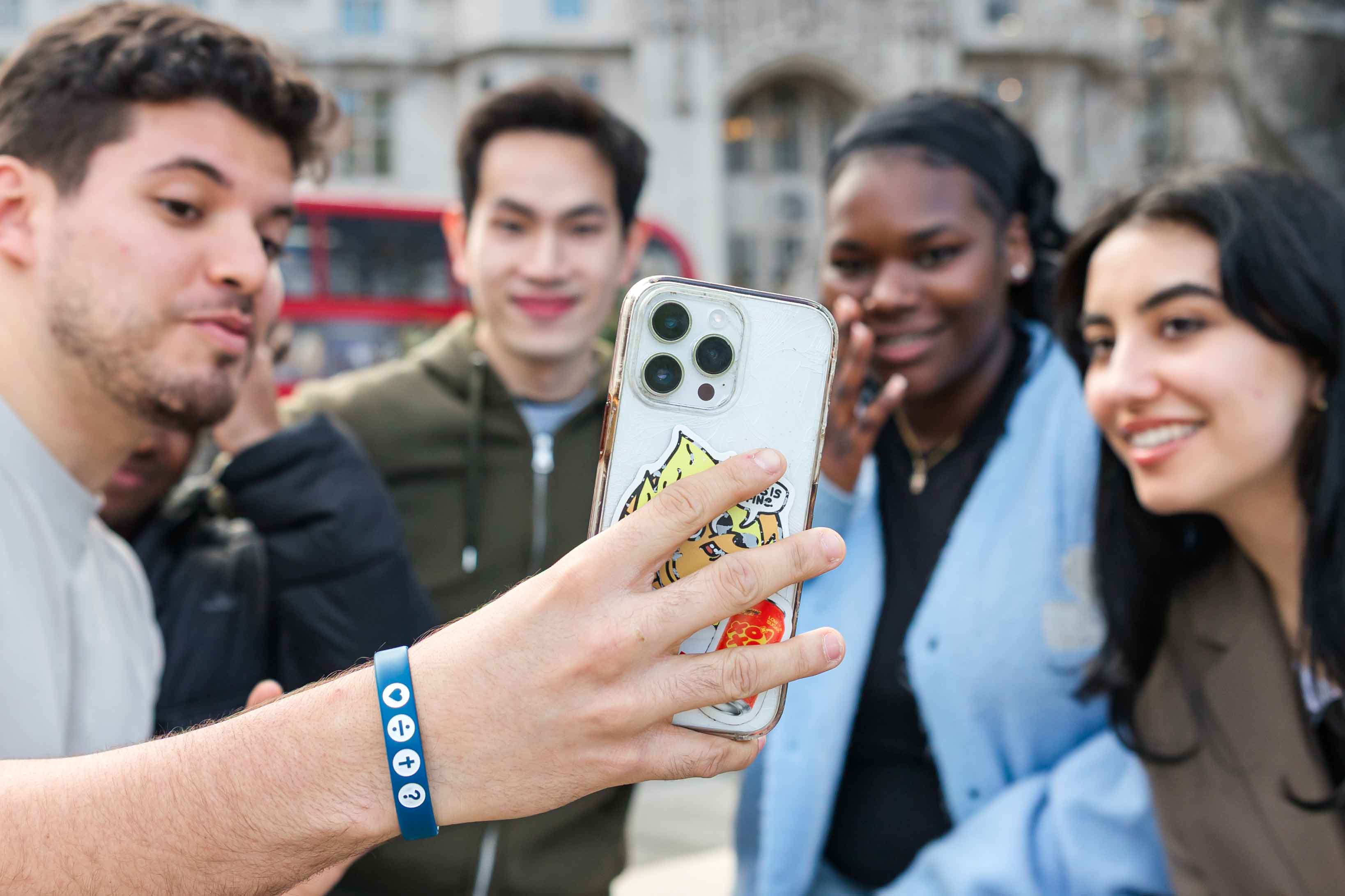A diverse group of four young adult friends smiling and waving while taking a phone selfie together outdoors in London.