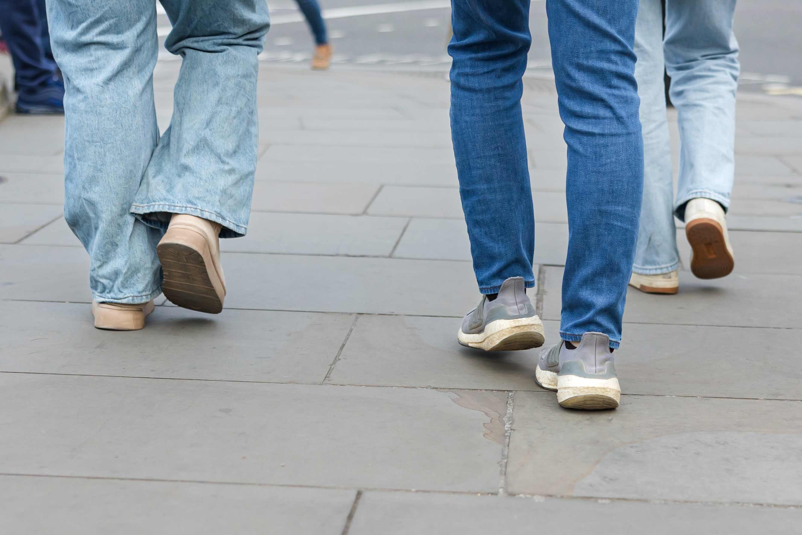 Close-up of the legs and feet of several people wearing jeans and casual shoes walking together on a paved sidewalk.