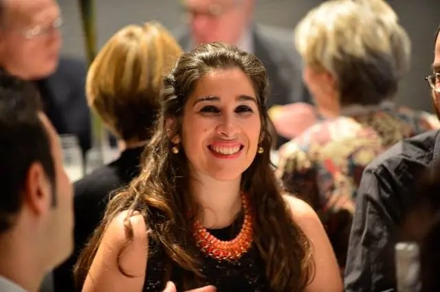 A smiling young woman with long dark hair, wearing a black top and an orange beaded necklace, socializing at a gathering or dinner event.
