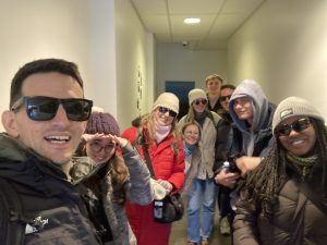 A group of nine people taking a selfie together in a brightly lit indoor hallway. They are dressed for cold weather in winter coats, beanies, and hoodies, with several members of the group wearing sunglasses indoors.