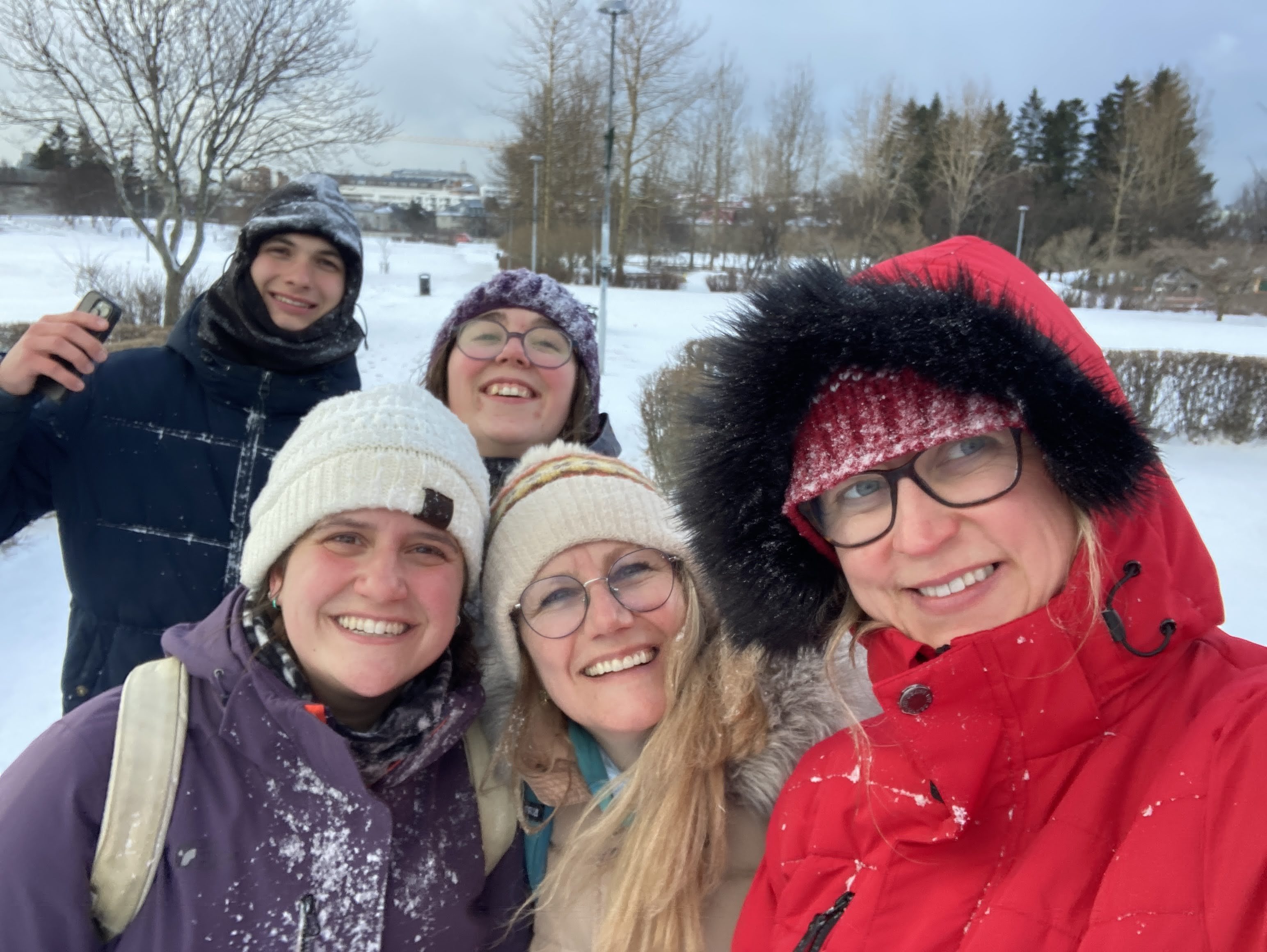 A smiling group of five people taking a selfie outdoors in a snowy, tree-lined landscape. They are bundled up in winter clothing, including heavy coats, knit beanies, and glasses, with specks of snow visible on their jackets.