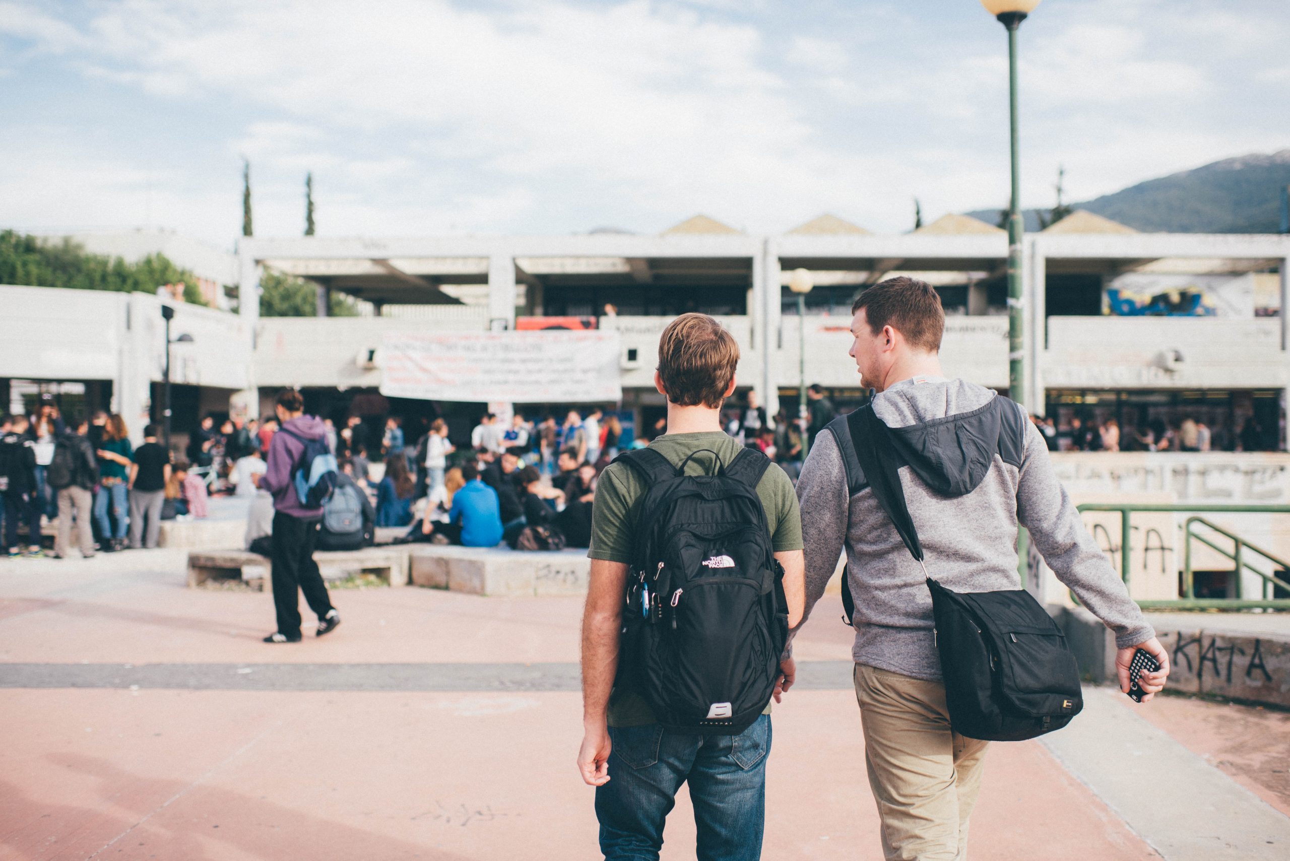 Two students with backpacks walking through a busy outdoor university campus or public square.