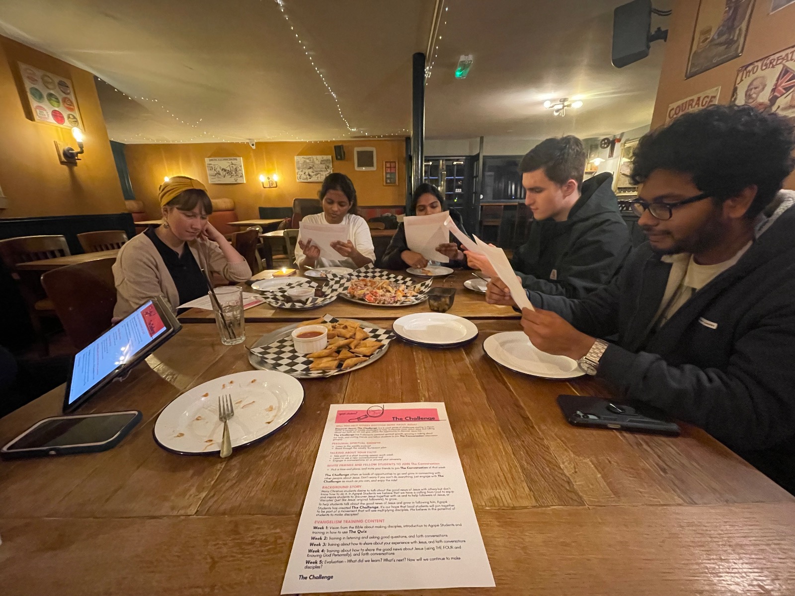 A group of young adults sitting around a wooden table in a cafe or pub, reading from discussion papers and sharing a meal together.
