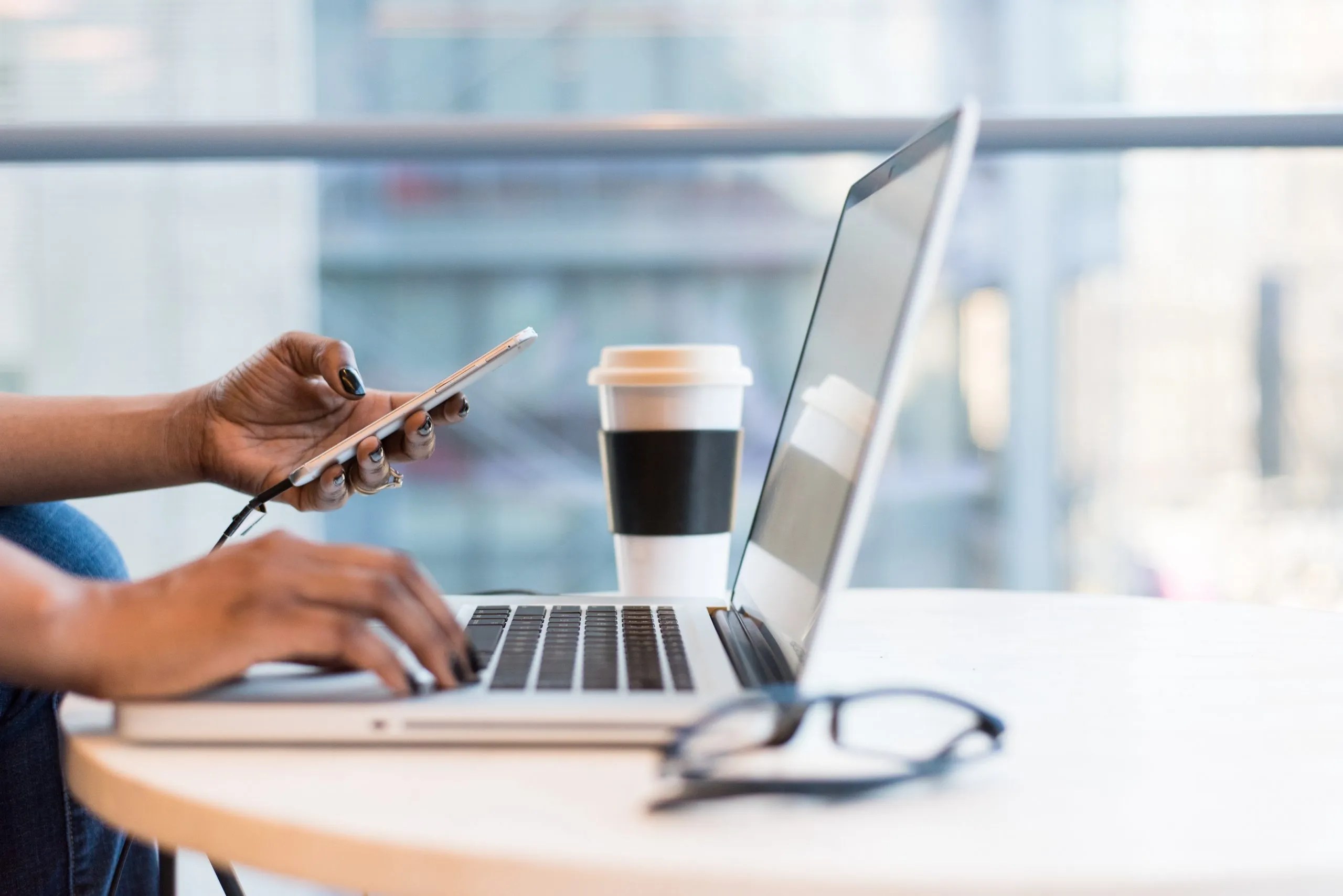 A person's hands holding a smartphone while typing on a silver laptop at a bright table with a paper coffee cup.