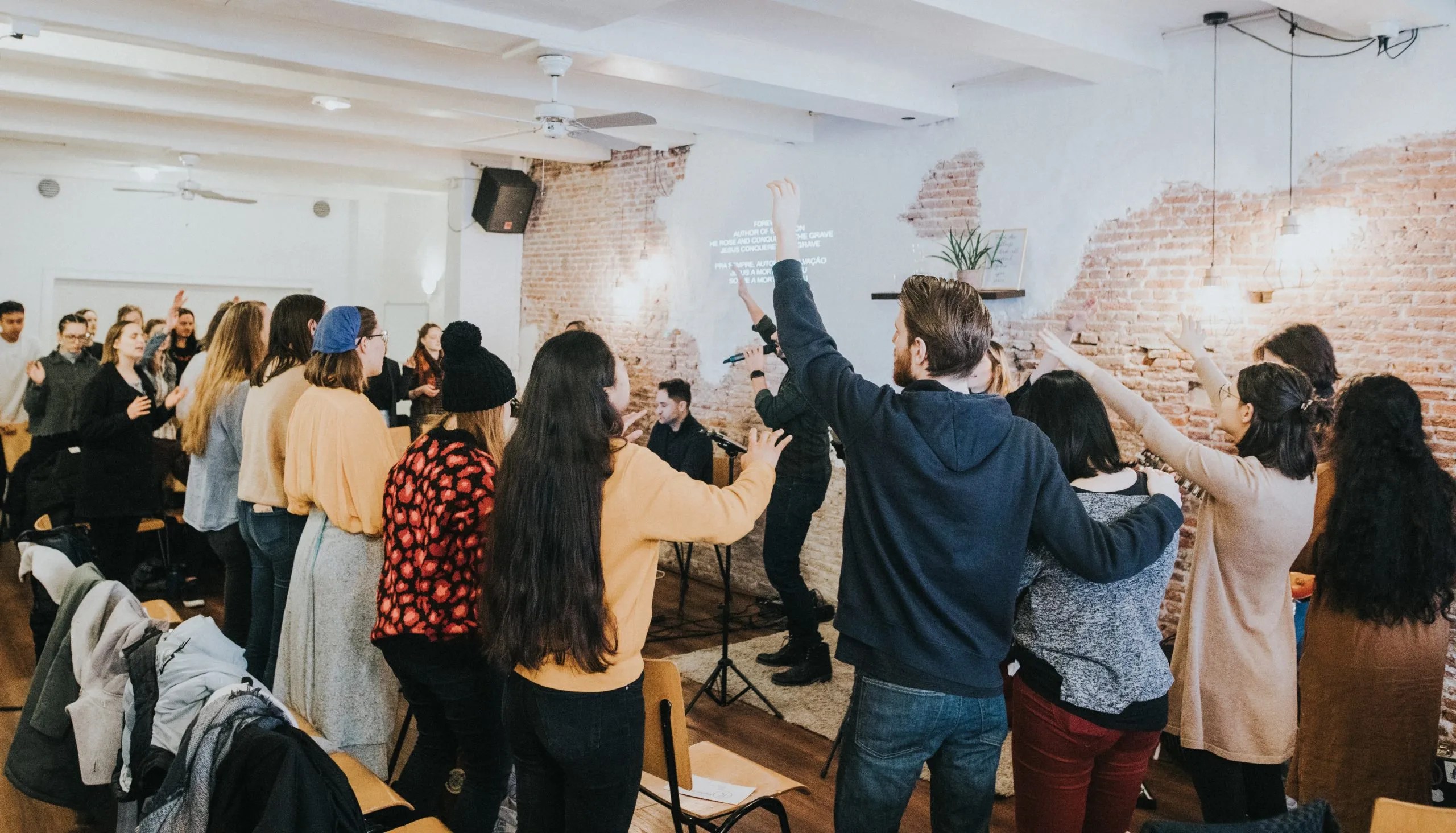 A diverse group of young adults standing in a room with exposed brick walls, lifting their hands in worship during a church service.
